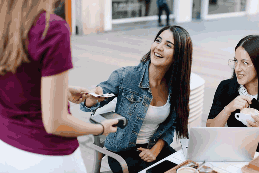 Foto de duas amigas em uma mesa. Uma delas está pagando a conta. Ao fazer isso, ela usa a matemática no dia a dia.