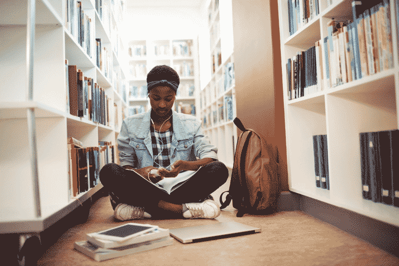 Foto de jovem negra sentada no chão de uma biblioteca, com livros e fones de ouvido. Aprender inglês com música é excelente para evoluir no idioma.