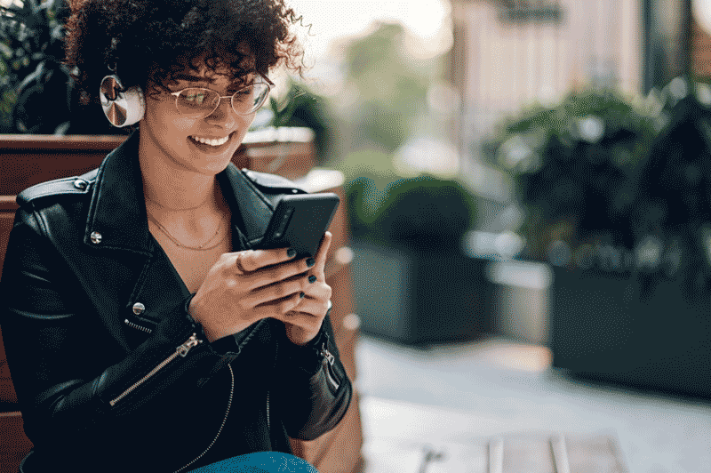 Foto de mulher jovem na rua, usando fones e mexendo no celular. É possível aprender inglês com música em qualquer lugar.