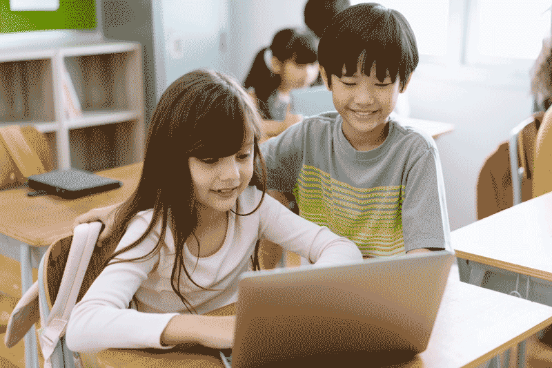 Foto de menina e menino sorridentes em sala de aula usando computador. A tecnologia na educação infantil é um ótimo modo de potencializar o conhecimento.