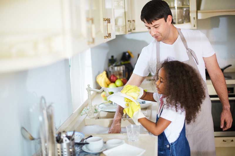 Foto de menina lavando louça junto com homem adulto. O papel dos pais é incentivar a autonomia infantil.