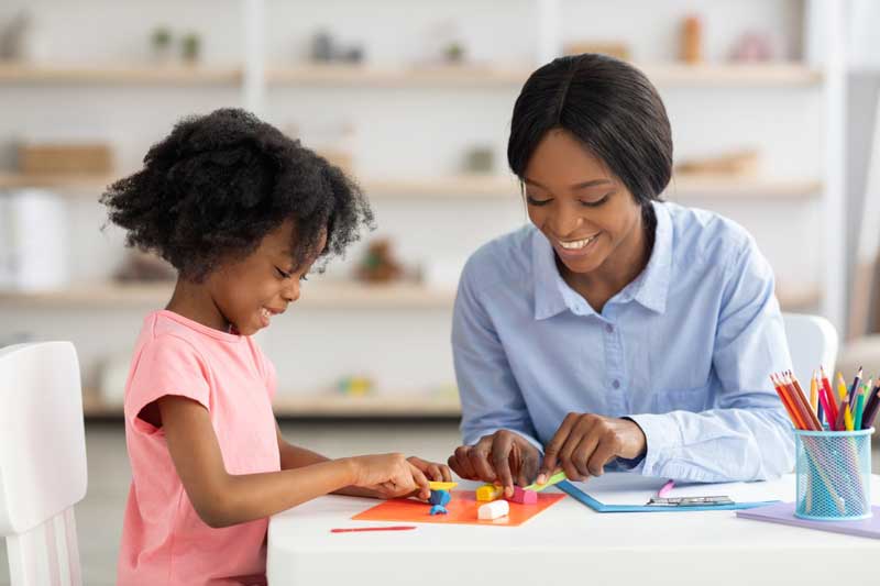 Foto de menina e mulher negras brincando sorridentes com massinha de modelar. Atividades para educação infantil como essa estimulam a criatividade.