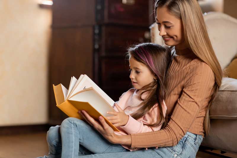 Foto de mãe e menina praticando o ensino bilíngue em casa, com a leitura de um livro.