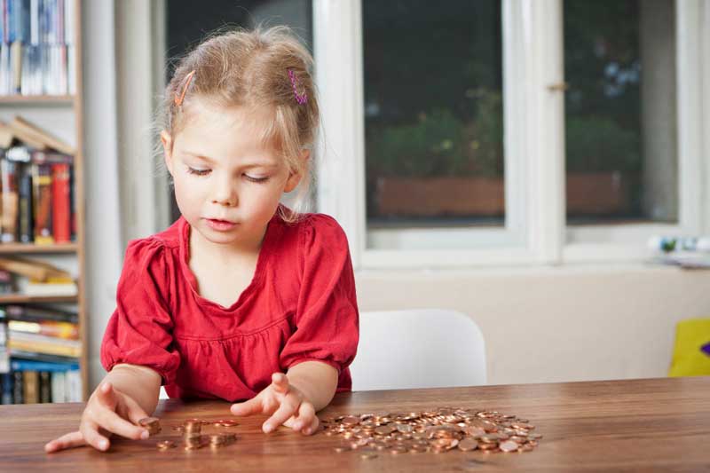 Foto de menina brincando com moedas sobre uma mesa. A educação financeira infantil começa com o exemplo dos pais.