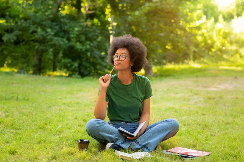 Foto de jovem mulher negra. Ela está sentada na grama, ao ar livre, pensativa. Segura um caderno e tem um lápis na mão. Estudar para o Enem exige muita dedicação.