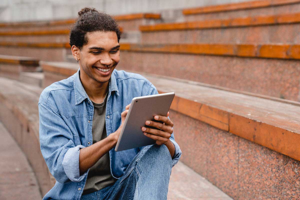 Foto de homem jovem sorrindo enquanto usa um tablet. Ele está sentado em um degrau ao ar livre. Usar a tecnologia para organização é um bom jeito de focar nos estudos.
