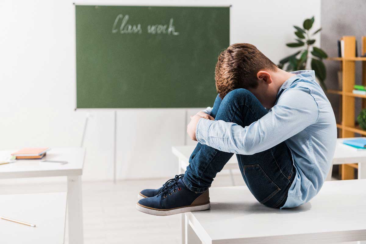 Foto de menino vítima de bullying na escola, sentado com a cabeça entre os joelhos, triste e isolado.
