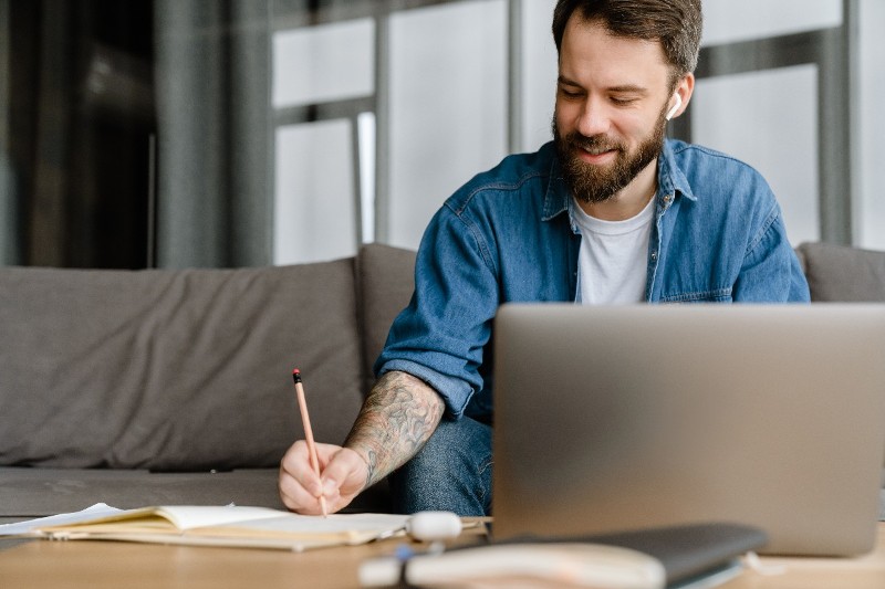 Foto de jovem homem branco com barba. Diante de um notebook, ele usa um lápis para escrever em um caderno. A repetição de palavras prejudica a coesão textual e deve ser evitada.