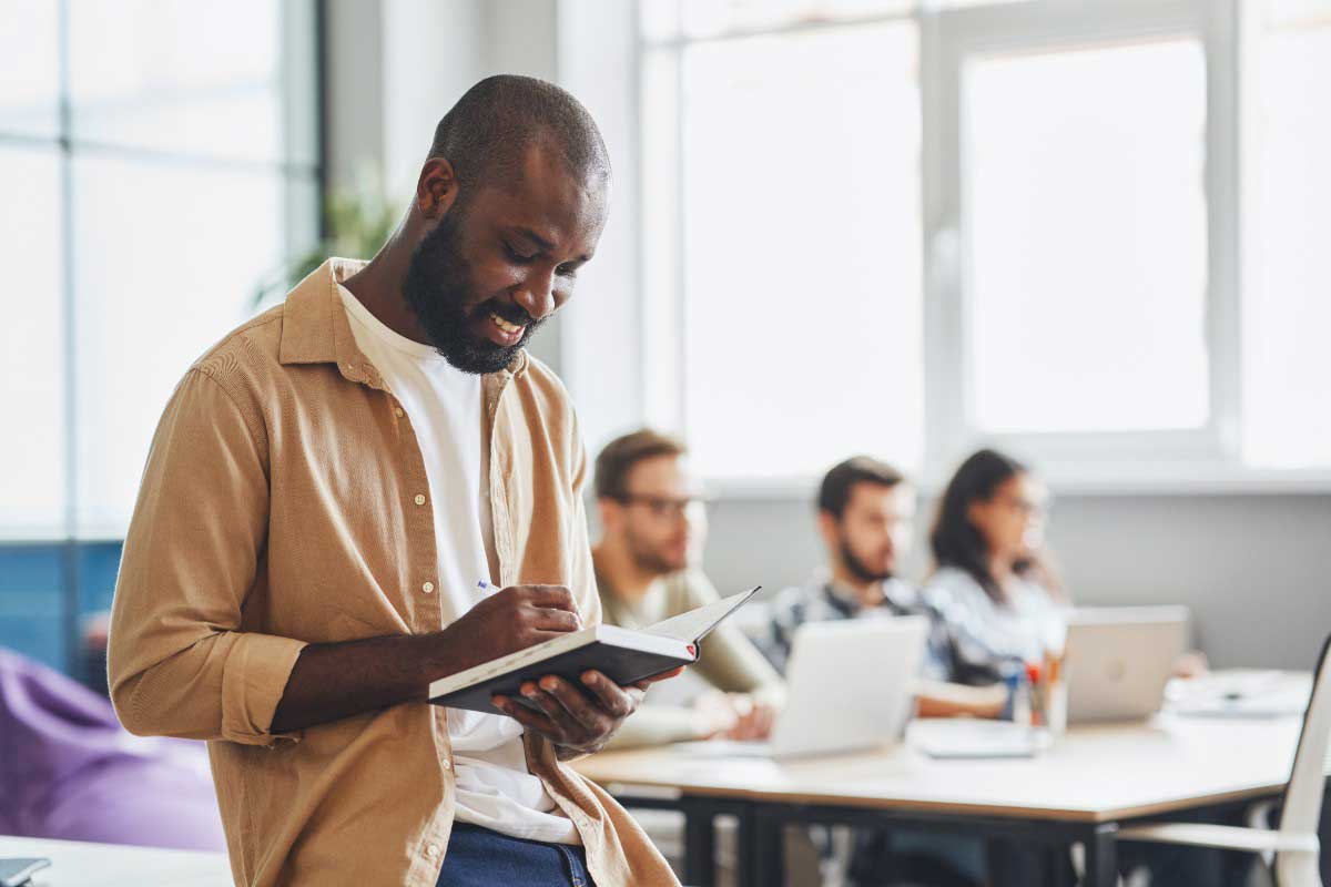 Foto de jovem negro em pé escrevendo em um caderno, como forma de treinar como melhorar o vocabulário.