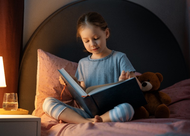Foto de menina sentada na cama lendo um livro.