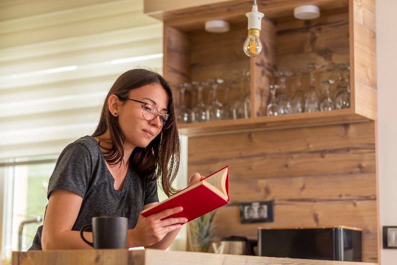 Foto de jovem mulher lendo um livro na mesa da cozinha.