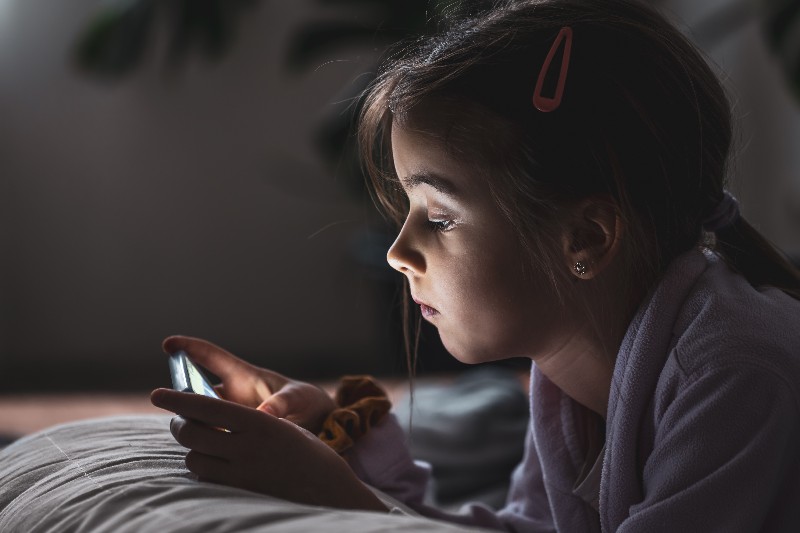 Foto de criança no celular em um quarto escuro, iluminado apenas pela tela do aparelho.