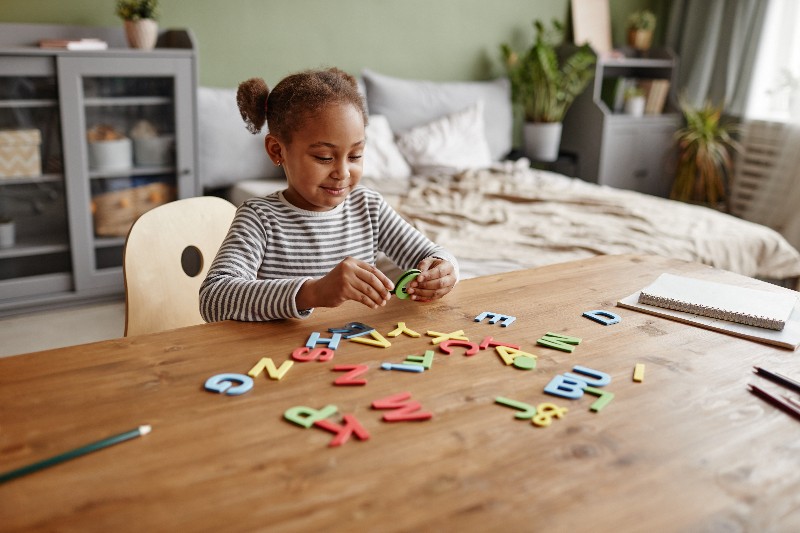 Foto de menina negra brincando com letras coloridas. Elas estão espalhadas sobre uma mesa, onde também há um caderno e lápis. No fundo há uma cama desarrumada.