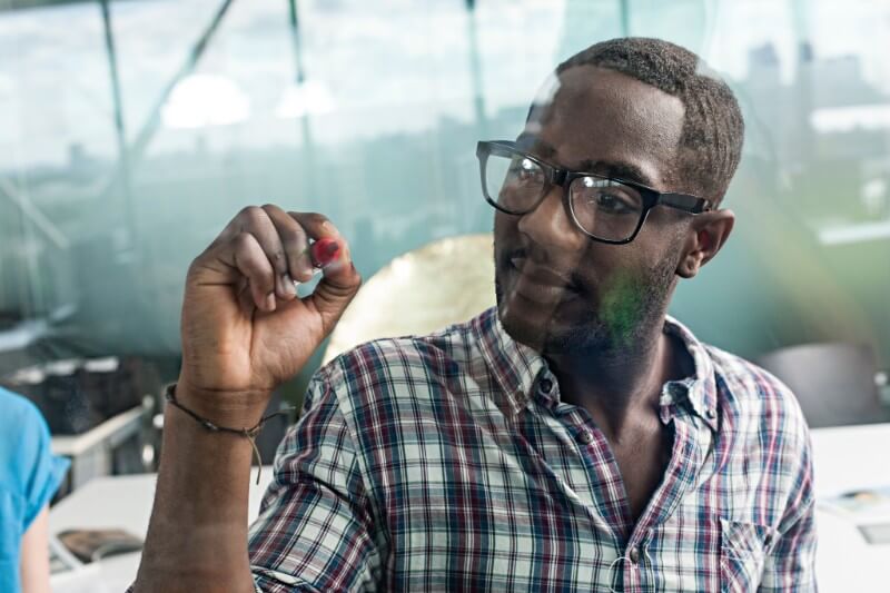 Foto de jovem homem negro treinando o uso de vírgula, com caneta vermelha, sobre um painel de vidro.