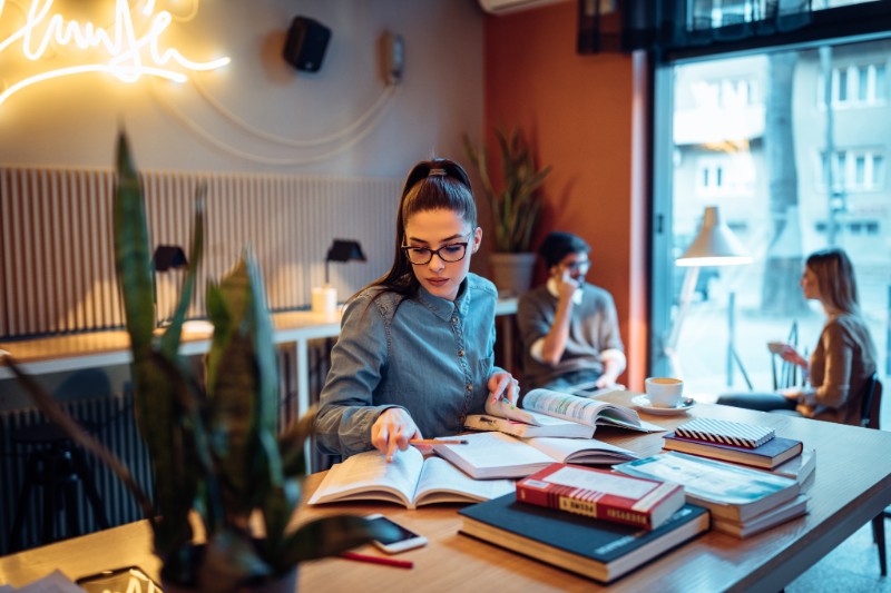 Foto de jovem mulher estudando em um café. Ela tem vários livros à mão..