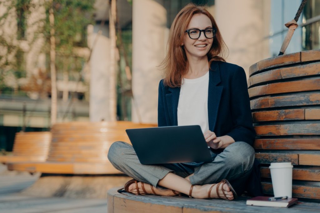 Foto de mulher ruiva sentada em um banco ao ar livre com um notebook no colo.