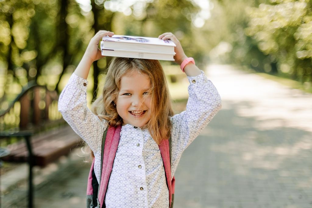 Menina sorridente indo para a escola com livros sob sua cabeça.
