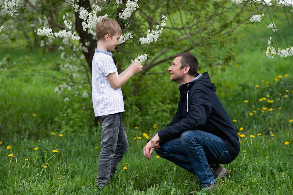 Pai sorridente conversando com um menino segurando uma flor branca em um jardim.
