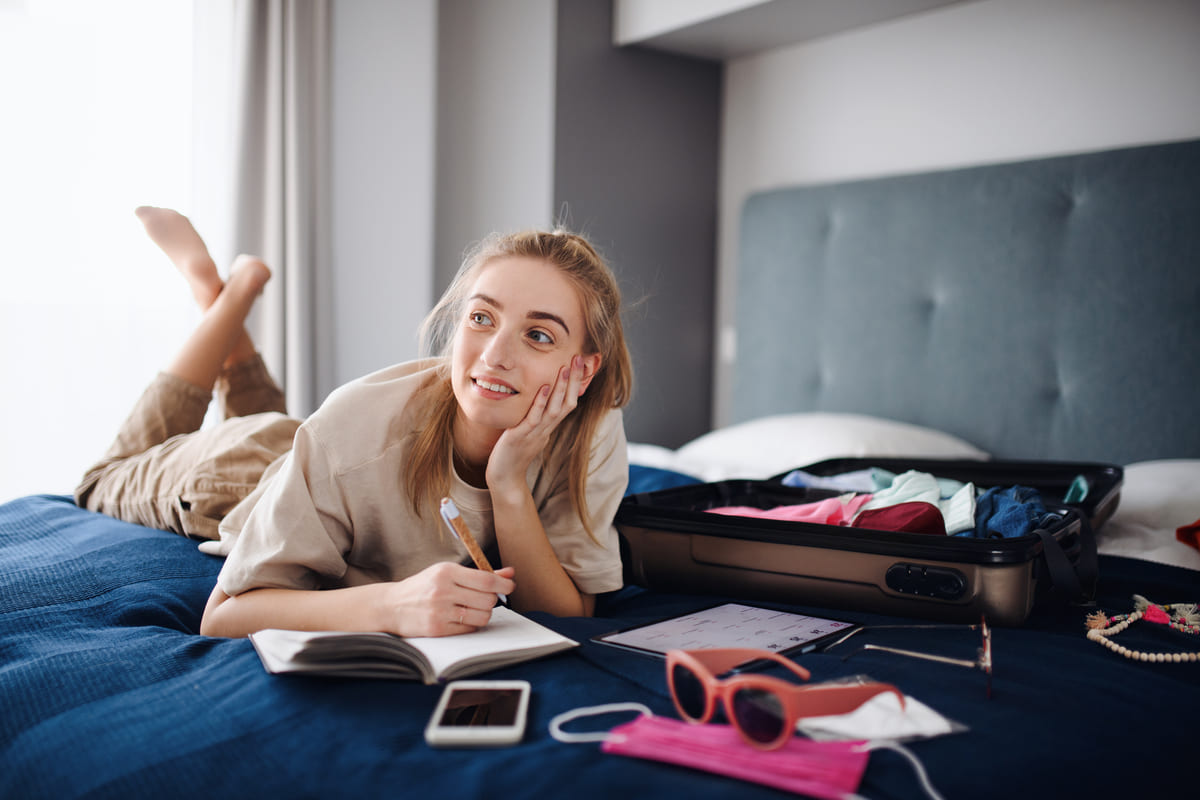 Jovem mulher escrevendo notas e embalando roupas na mala, planejando primeira viagem internacional.