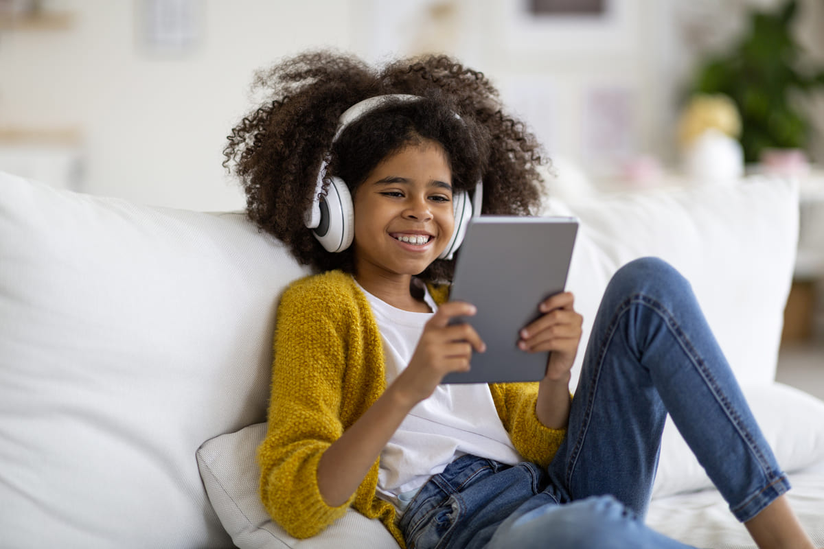 Criança feliz sentada no sofá, usando tablet digital e fone de ouvido.