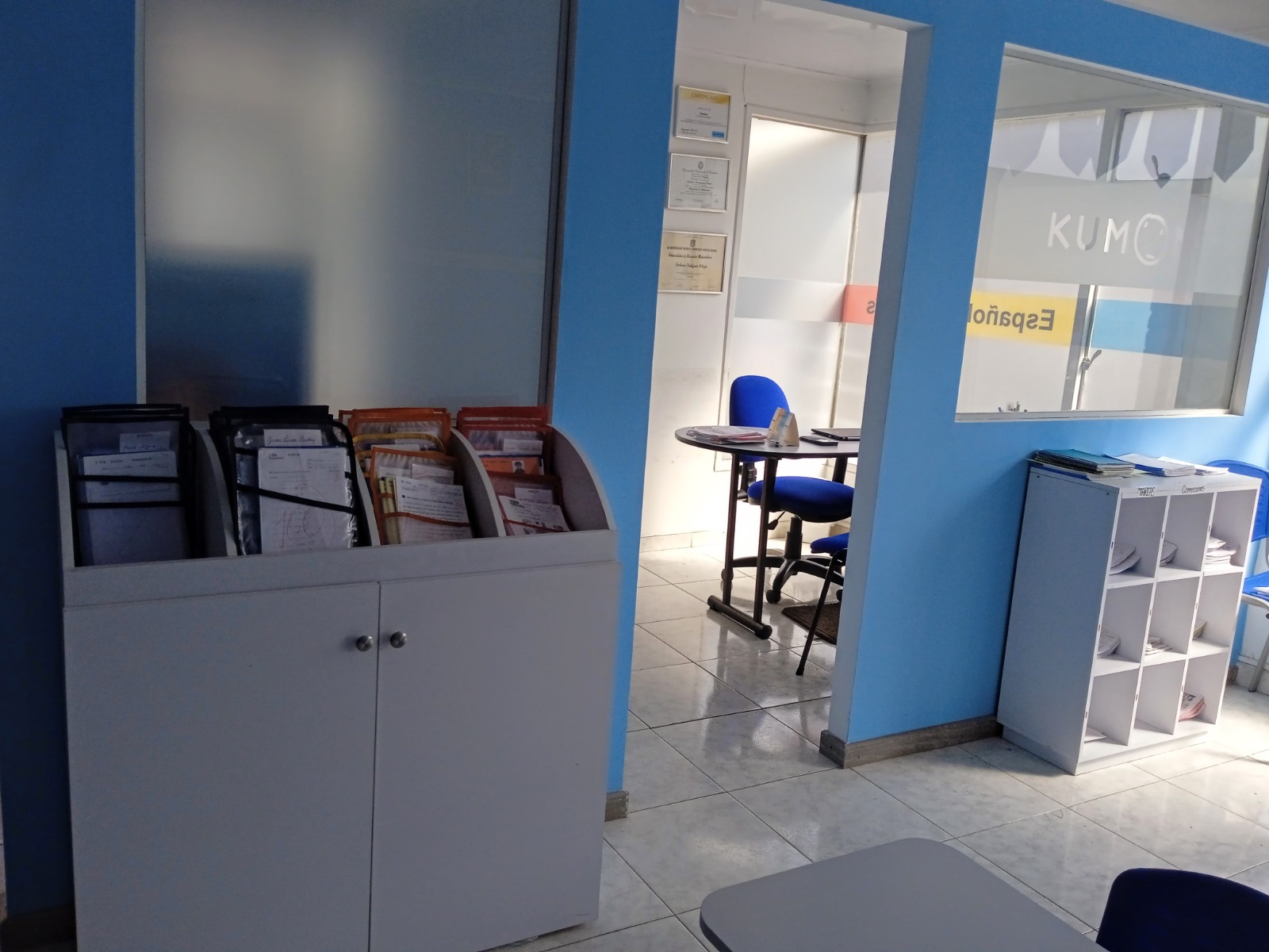 Interior de la sala de clases de Kumon en Pasadena, con mesas blancas y sillas azules, y un gran cuadro de avance de alumnos en la pared.