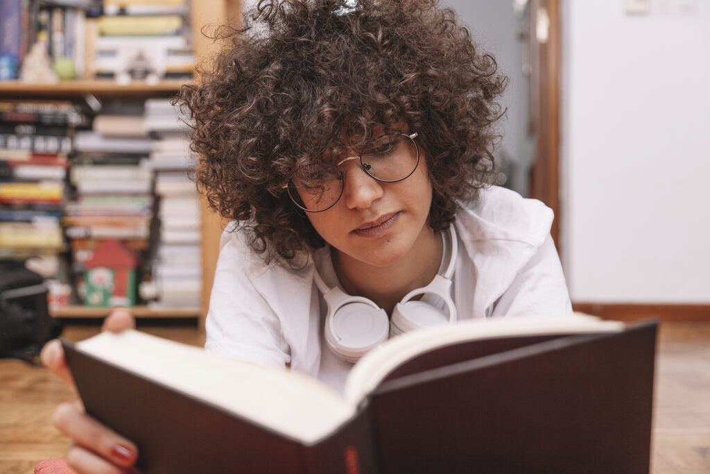 Adolescente concentrada lendo livro, com estante repleta de livros no fundo, representando a importância da leitura como hábito para aprender como escrever uma redação clara e coerente.