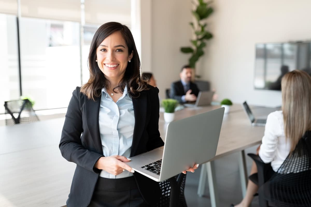 Empreendedora sorridente segurando notebook com ambas as mãos, enquanto colaboradores fazem reunião atrás.