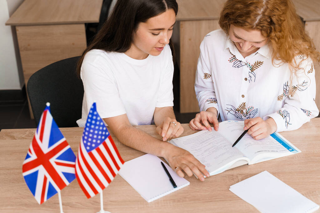 Duas mulheres estudando inglês com livros em cima de mesa, e duas bandeiras de países anglófonos ao lado.