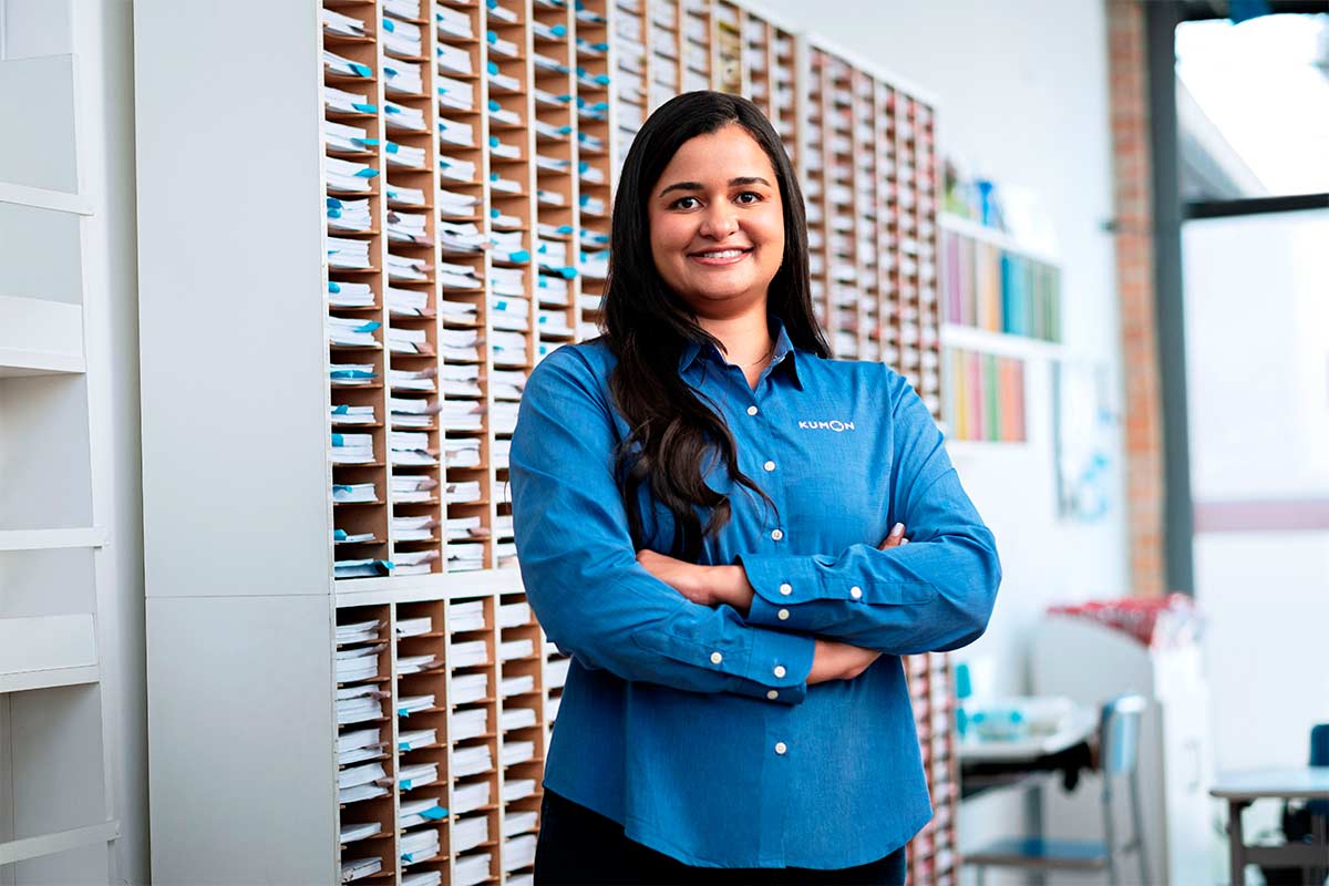 Mulher sorrindo com os braços cruzados posando para a foto dentro de unidade do Kumon